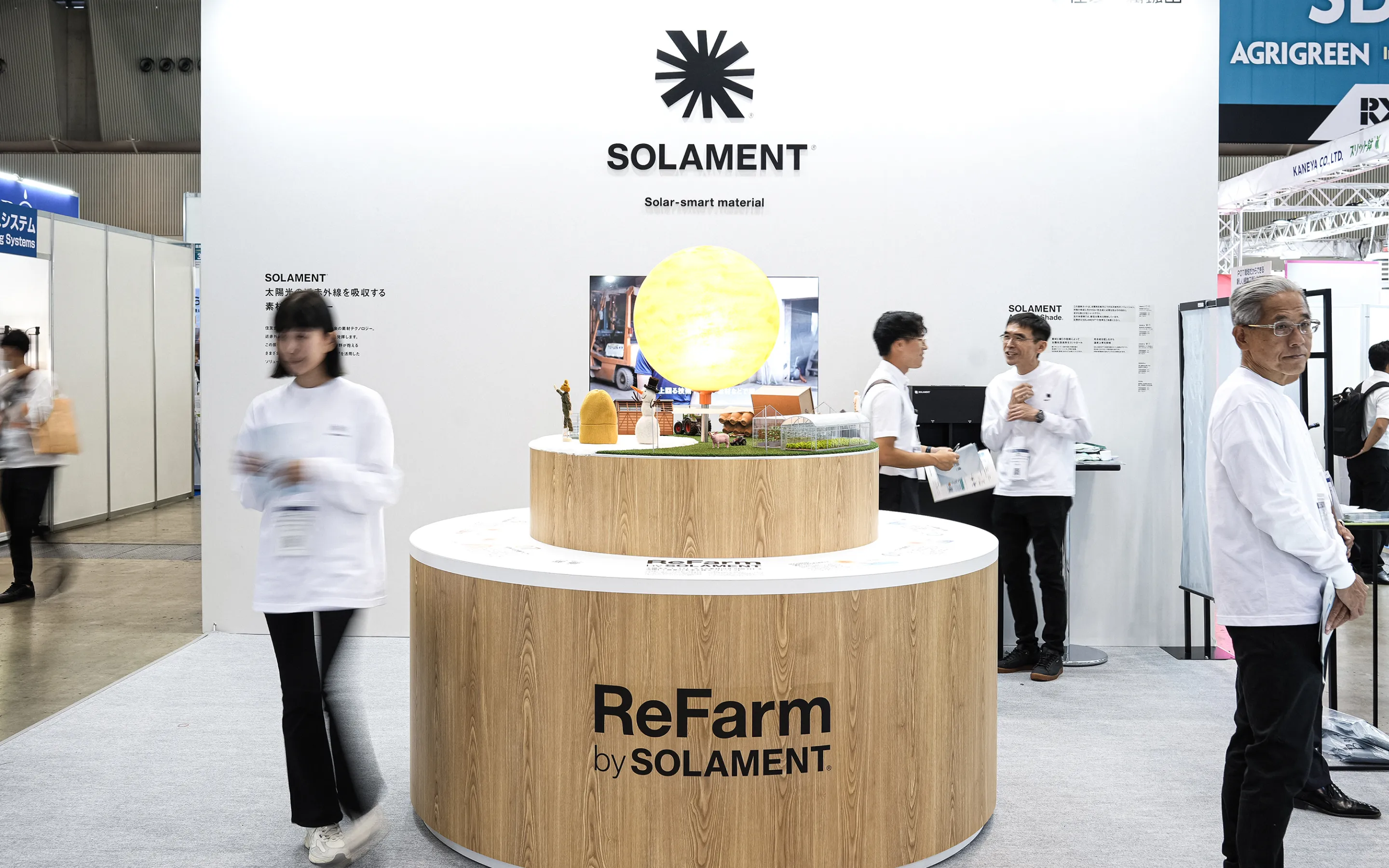 農業WEEKの様子。中央にはReFarm by SOLAMENT®の木目調の円柱形展示台。上段には光る太陽と農場のジオラマが飾られ、周囲にスタッフや来場者がいるビジュアル
