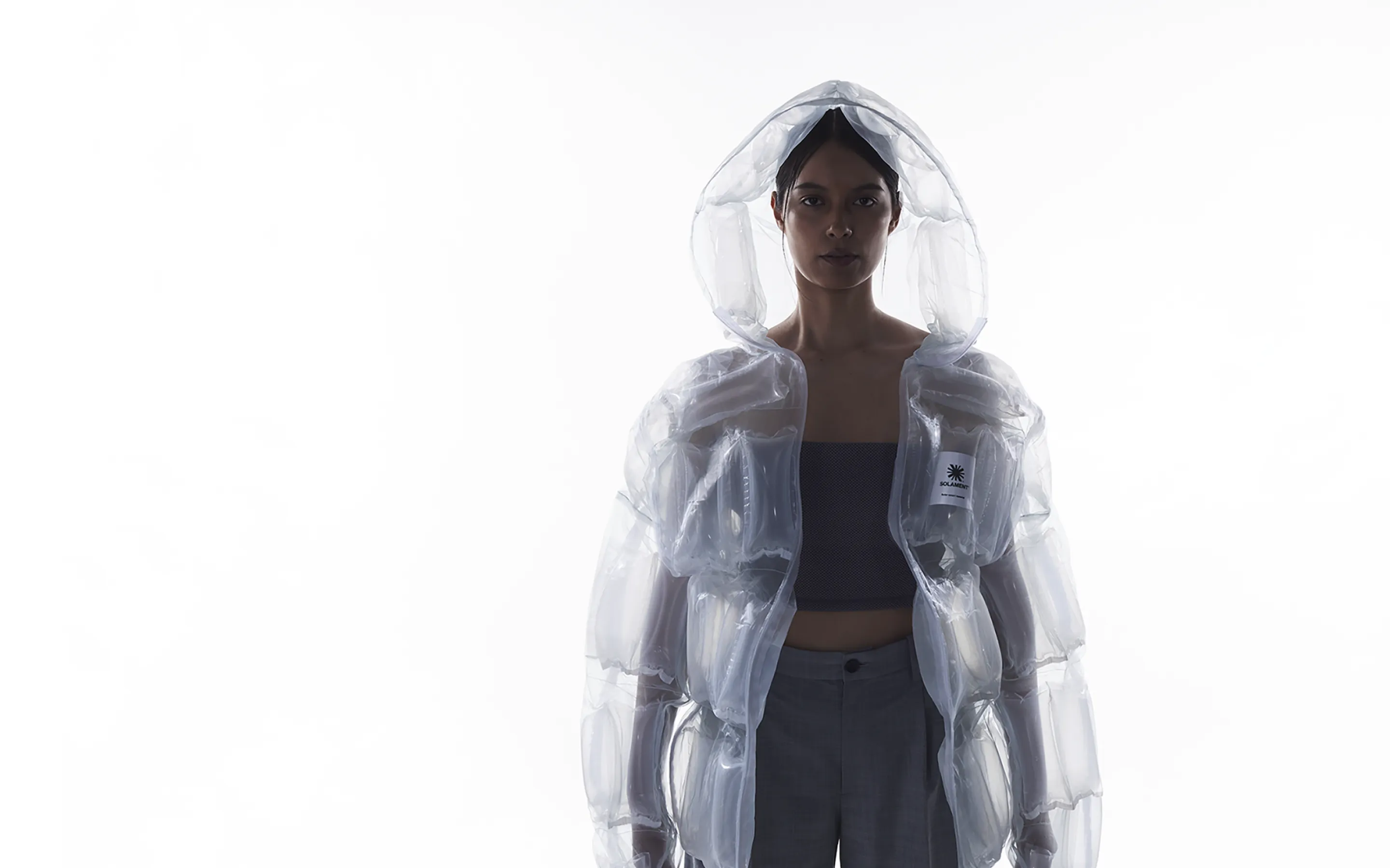 透明素材のフード付きジャケット（DOWN-LESS DOWN JACKET）を着用した女性。空気の入った構造が「少ない資源で圧倒的な機能性」という特徴を表現。