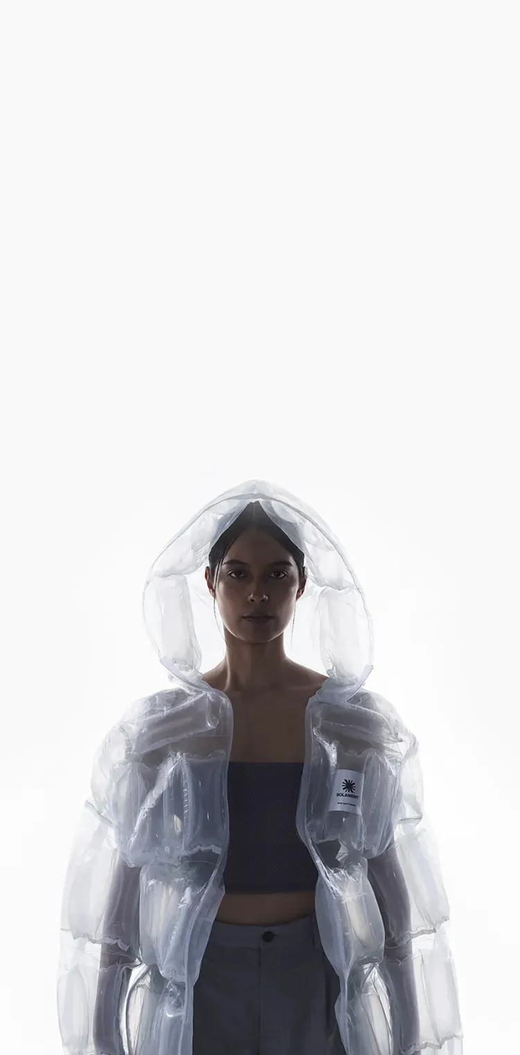 透明素材のフード付きジャケット（DOWN-LESS DOWN JACKET）を着用した女性。空気の入った構造が「少ない資源で圧倒的な機能性」という特徴を表現。