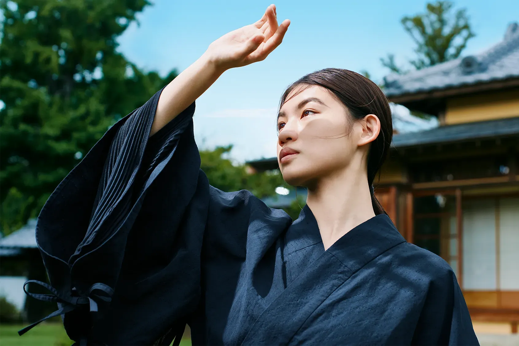 日本家屋を背景にHOPE WEAR for Cultureを着た女性。手を掲げ、大きな袖の内側にあるプリーツ加工の通気性を示している。