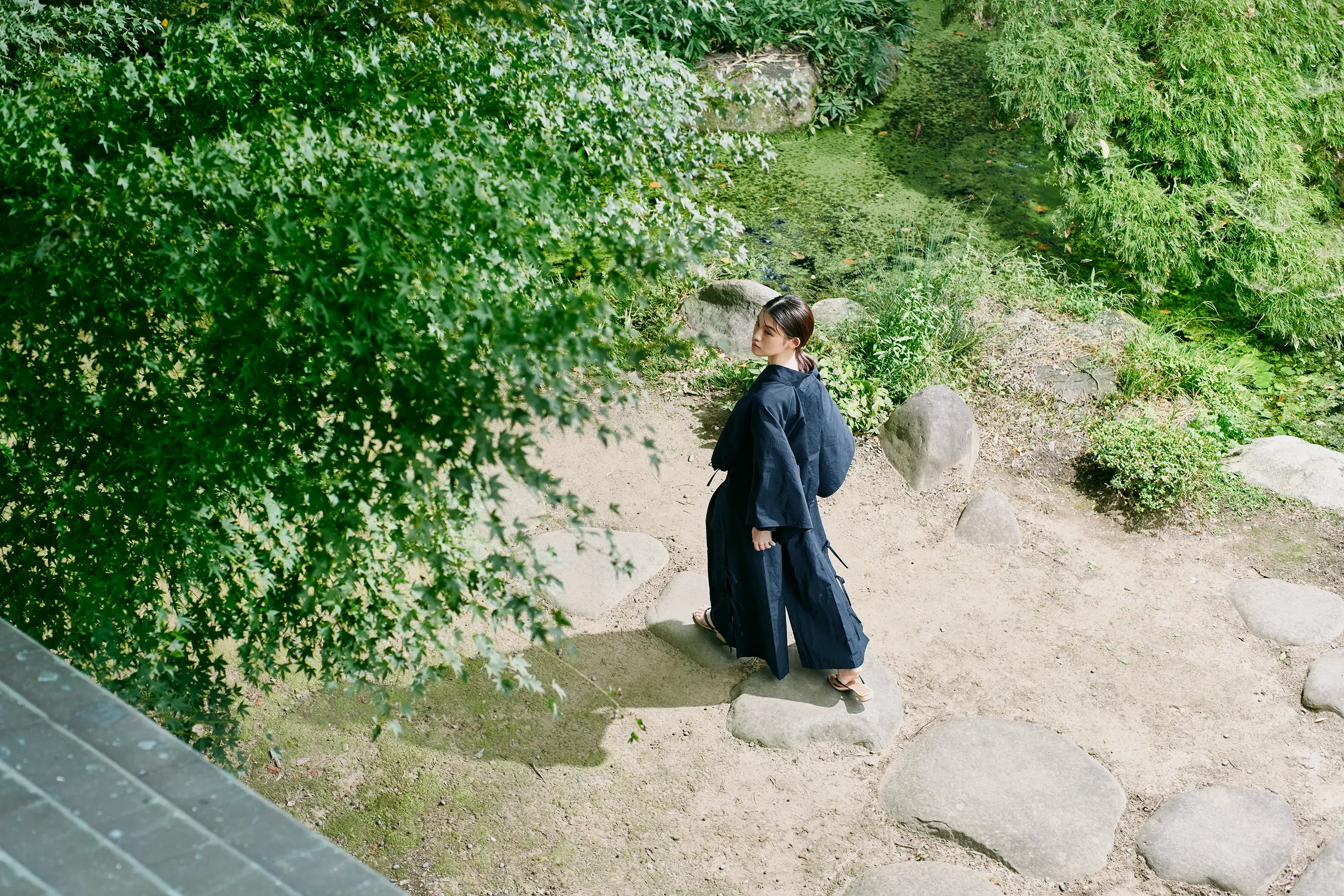 日本庭園の飛石を歩く女性。濃紺の和服風セットアップ（HOPE WEAR for Culture）の大きな袖が広がり、夏の快適性を表現している。
