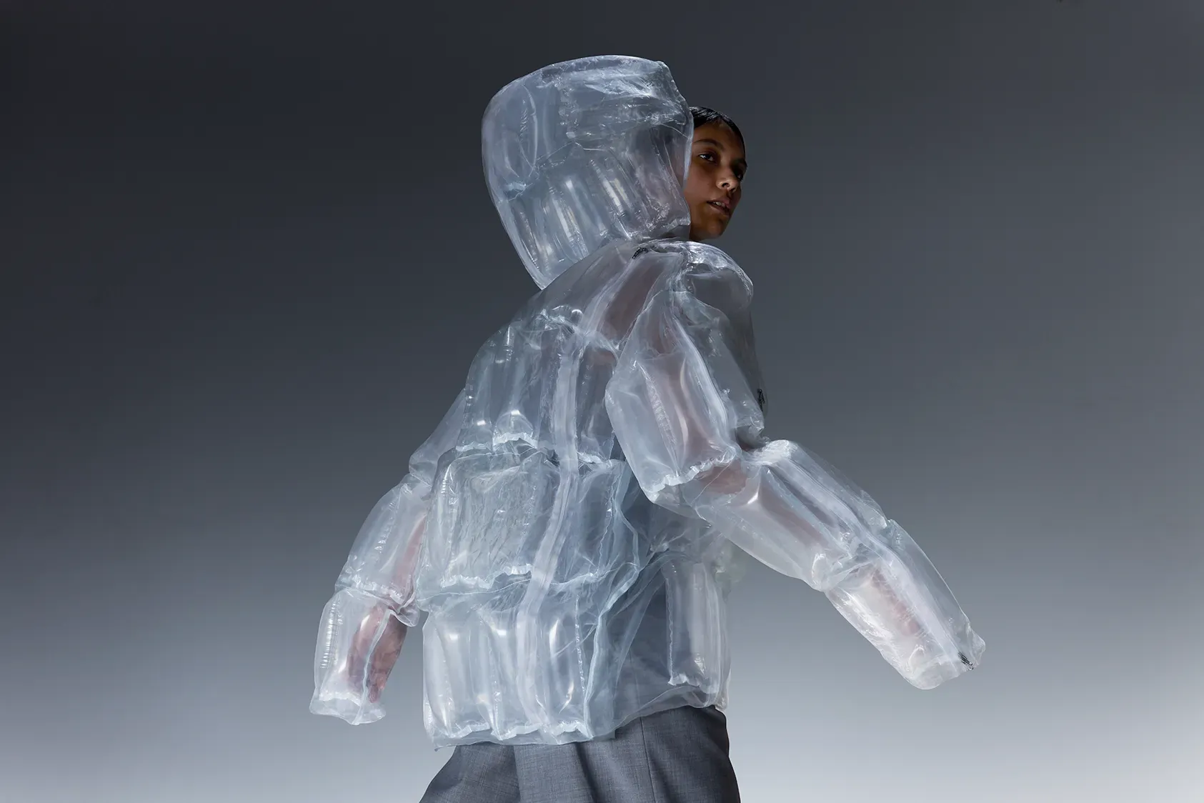 透明なDOWN-LESS DOWN JACKETを着て振り返るモデル。羽毛を一切使用せず、空気の層で構成された太陽光発熱ダウンジャケット。