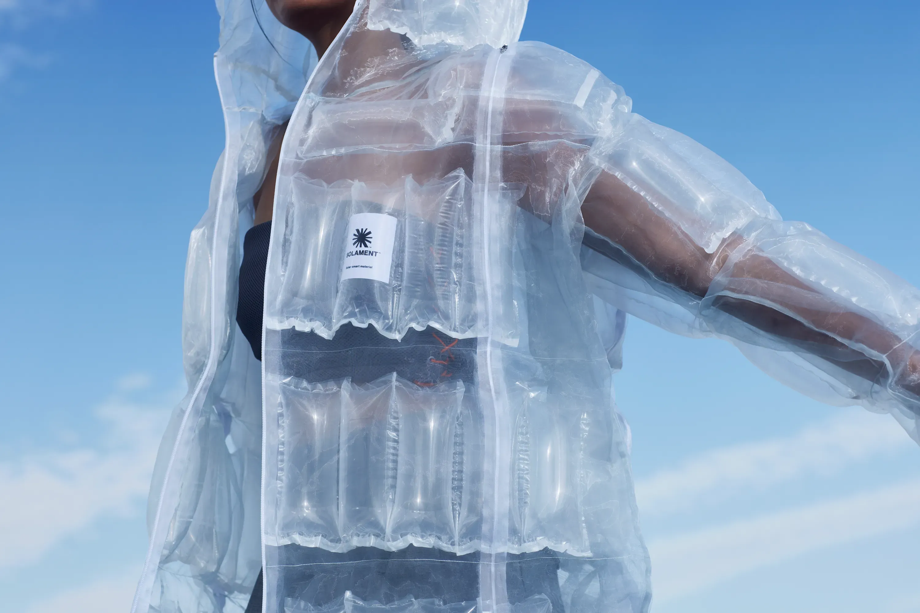 青空の下、透明なDOWN-LESS DOWN JACKETを着たモデル。太陽光で発熱する空気のブロックとタグが透けて見える。