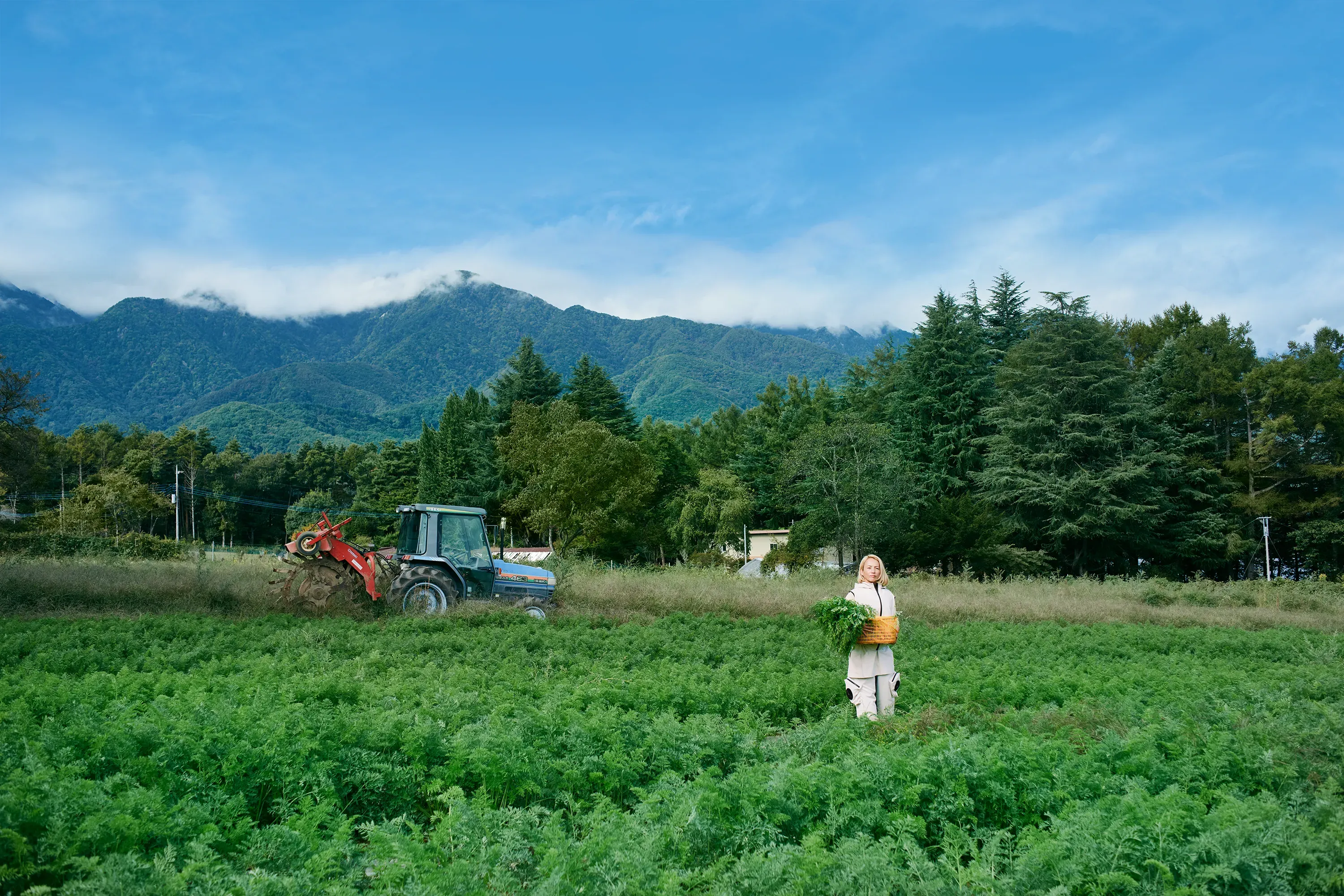 農場を背景に、HOPE WEAR for Farm Type B（ベストとエプロン付きパンツ）を着て野菜カゴを持つ女性。左奥でトラクターが稼働中。